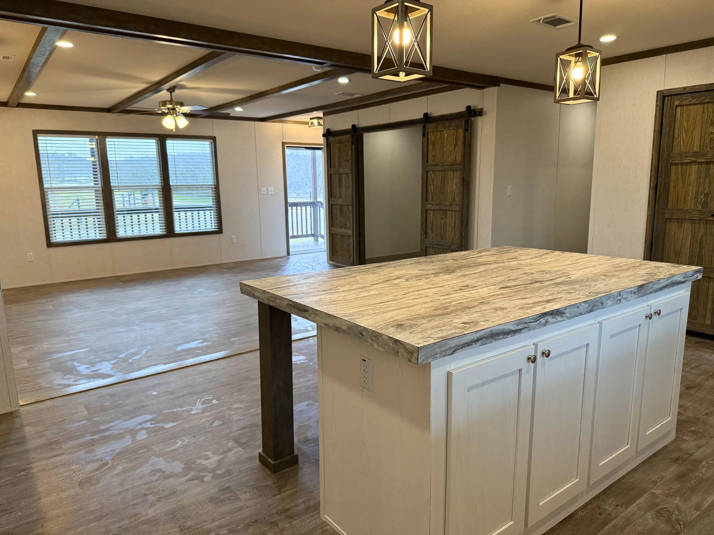 Empty living room and kitchen area with large windows, wooden sliding barn doors, a kitchen island with a wood countertop, and pendant lights, in a modern home.