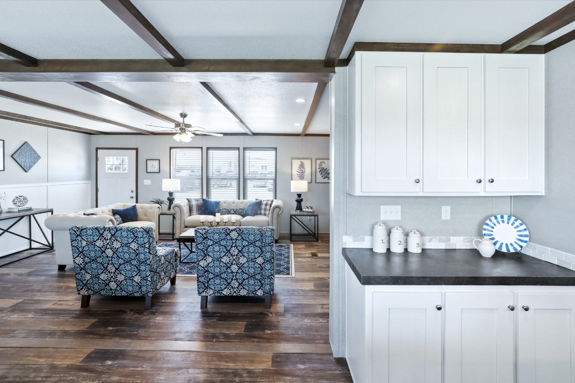 Living room with white sofas, patterned blue armchairs, wooden floors, large windows, and decorative wall art. Part of the kitchen with white cabinets and black countertop is visible in the foreground.