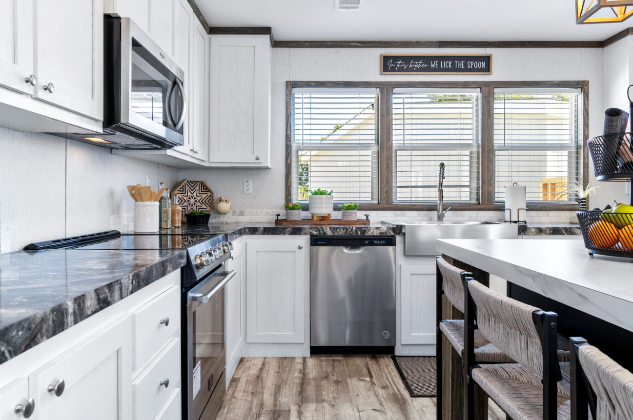Modern kitchen with white cabinets, stainless steel appliances, and wooden accents. A window above the sink provides natural light. There are decorative items and a sign that says 'In this kitchen we lick the spoon'.