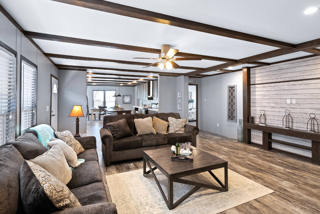 Living room with dark brown sofas, beige throw pillows, a wooden coffee table, a cream-colored rug, wooden floor, and a wall with light-colored shiplap paneling. In the background, a dining area and kitchen are visible, with a ceiling fan overhead an