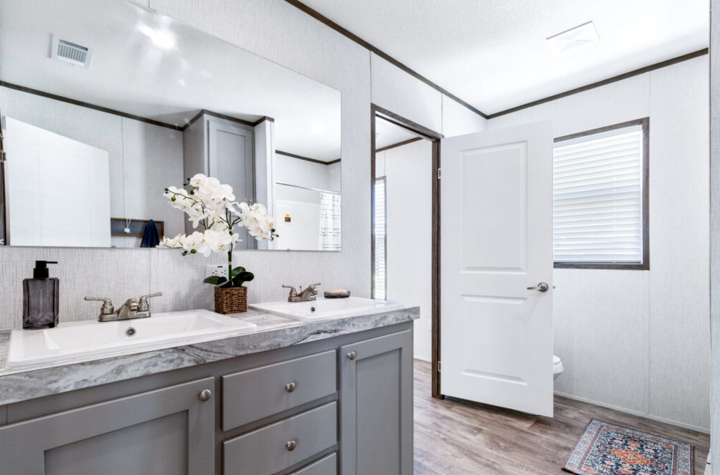 Bathroom with double vanity, gray cabinets, marble countertop, large mirror, orchid plant, soap dispenser, window with blinds, door, and a small rug on wood floor.