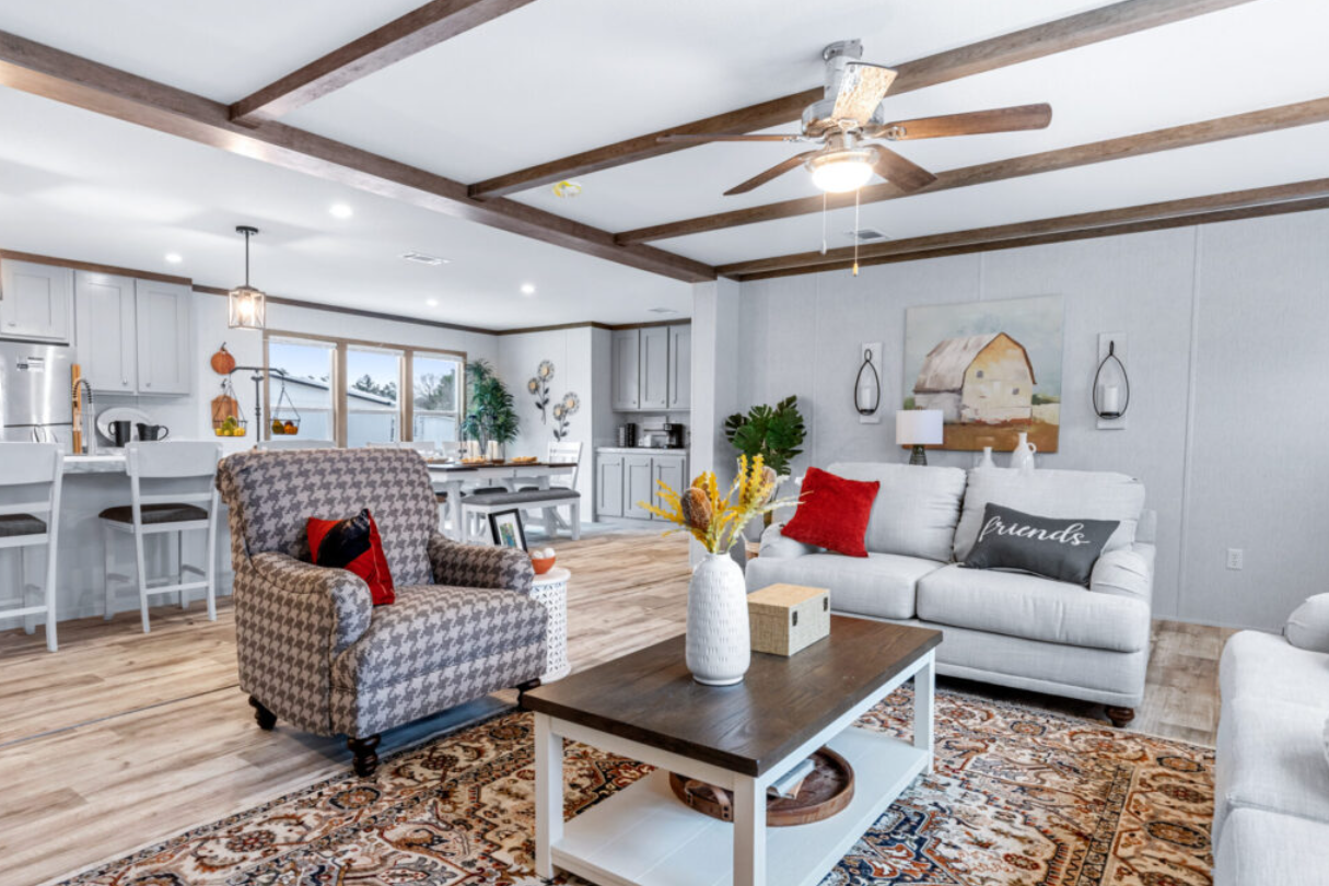 Open-concept living room and kitchen with wood beams on ceiling, white walls, hardwood floors, white sofa with red and black pillows, checkered armchair, wooden coffee table with a vase of flowers, and kitchen area with bar stools.
