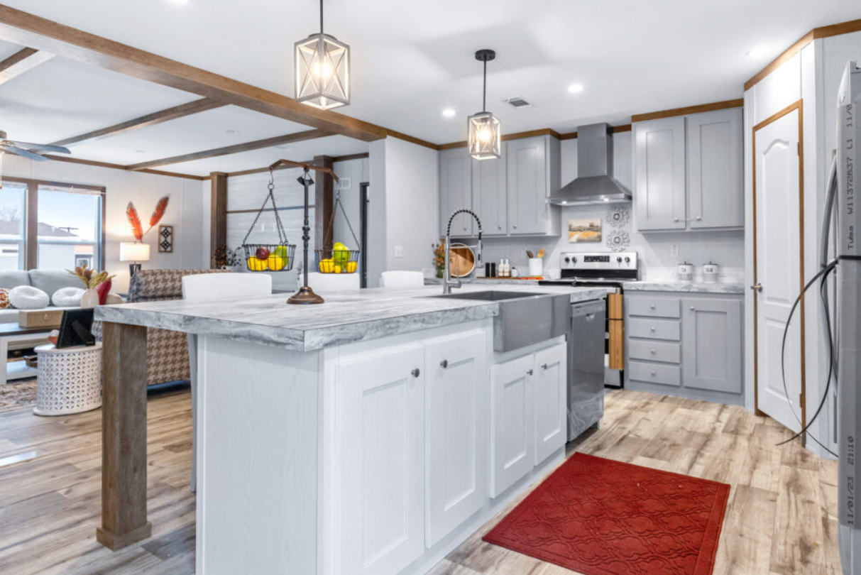 Modern kitchen with light gray cabinets, stainless steel appliances, and a white marble island. Decor includes a red rug, two hanging pendant lights, and a basket of fruit. Open living space with seating area and large windows.