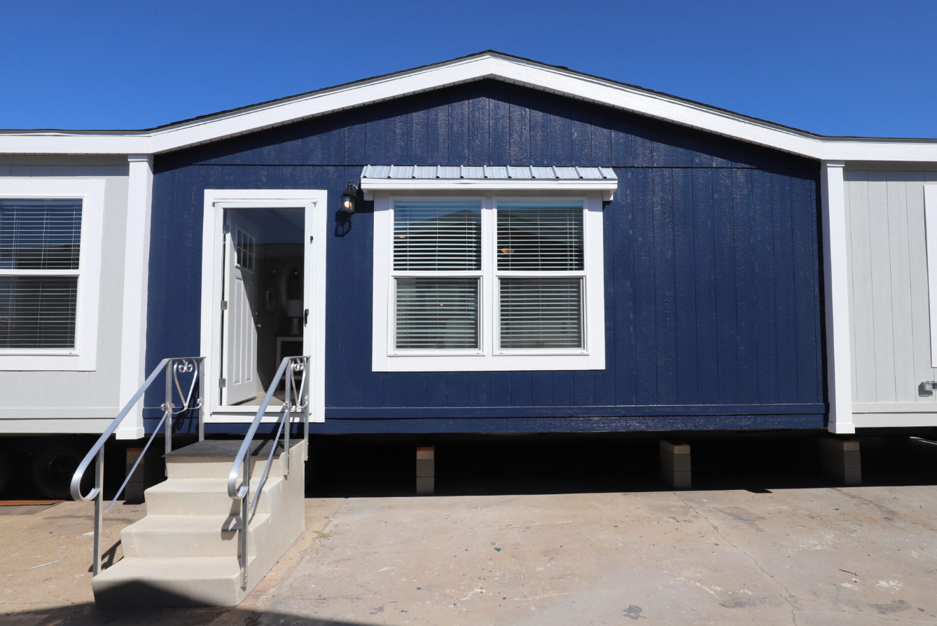 View of a tiny house with a dark blue exterior, white trim, a front door, and a window with blinds, on a concrete lot under a clear blue sky.