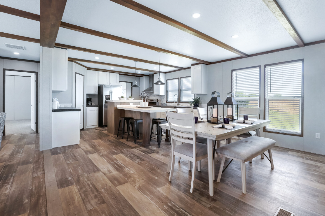 Open-concept kitchen and dining area with white cabinetry, stainless steel appliances, wooden beams, and a wooden floor. Dining table with four chairs and decorative lanterns near windows.
