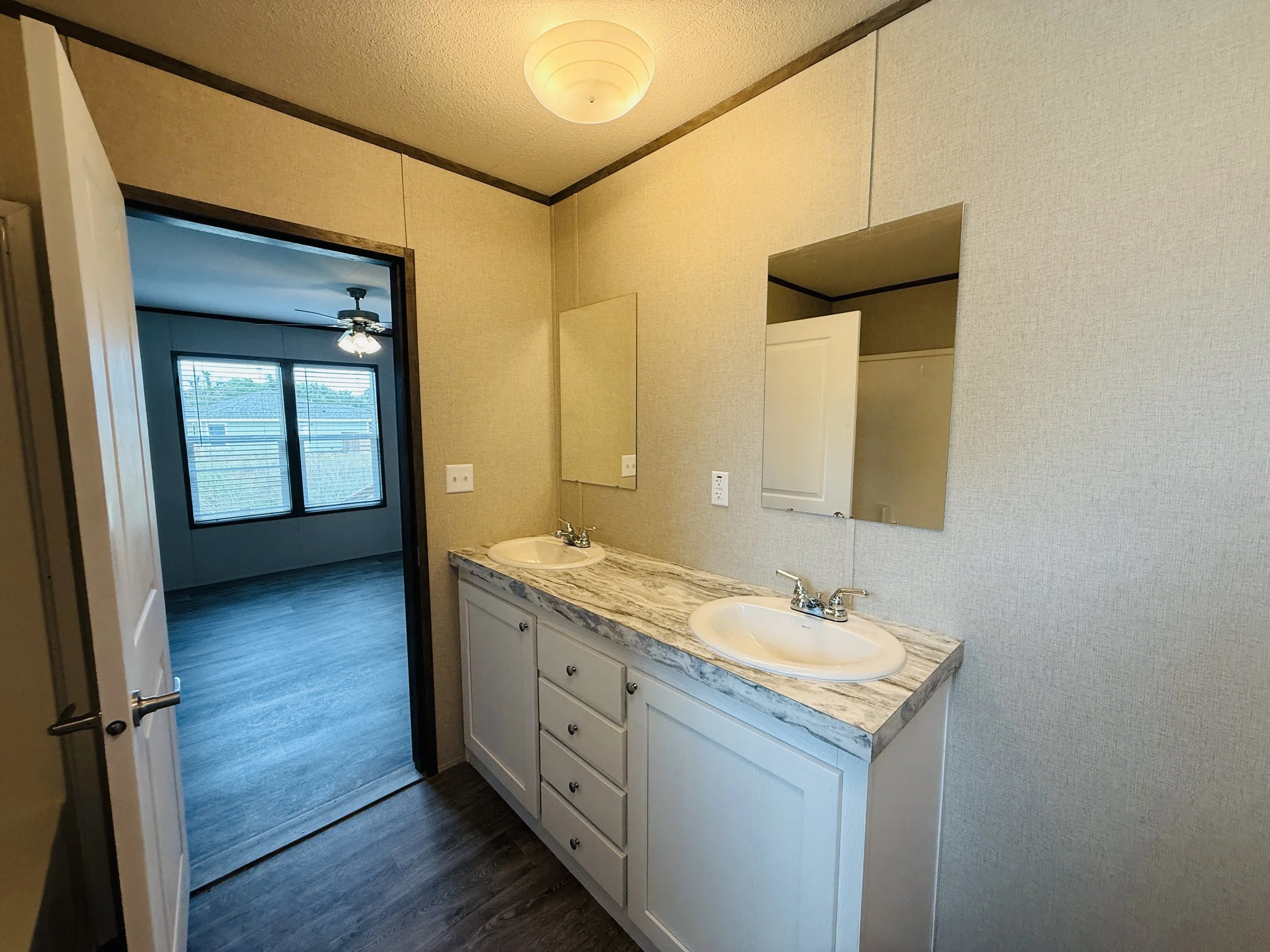 Bathroom with dual sinks, marble countertop, two mirrors, beige wallpaper, and a doorway leading to a room with large window and ceiling fan.