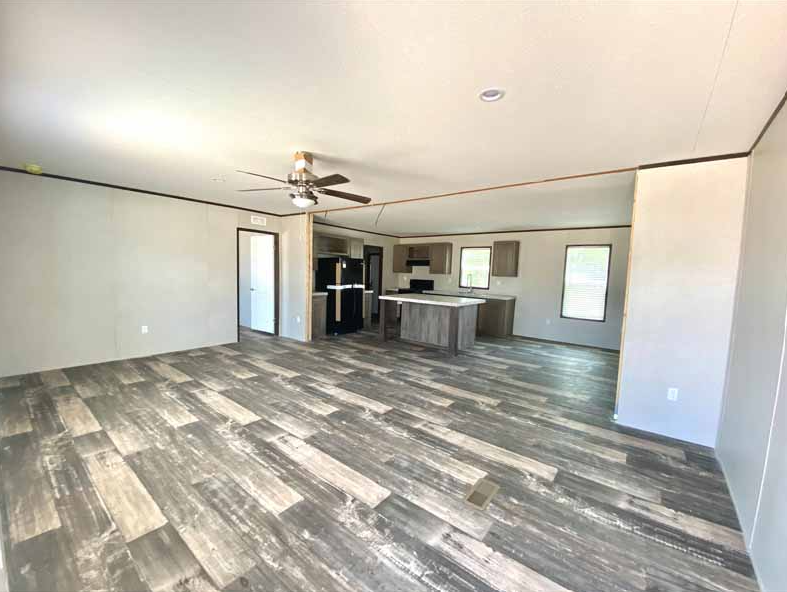 Empty living room with wood-look flooring, ceiling fan, and an open kitchen area with island and cabinets, three windows, and plain walls.