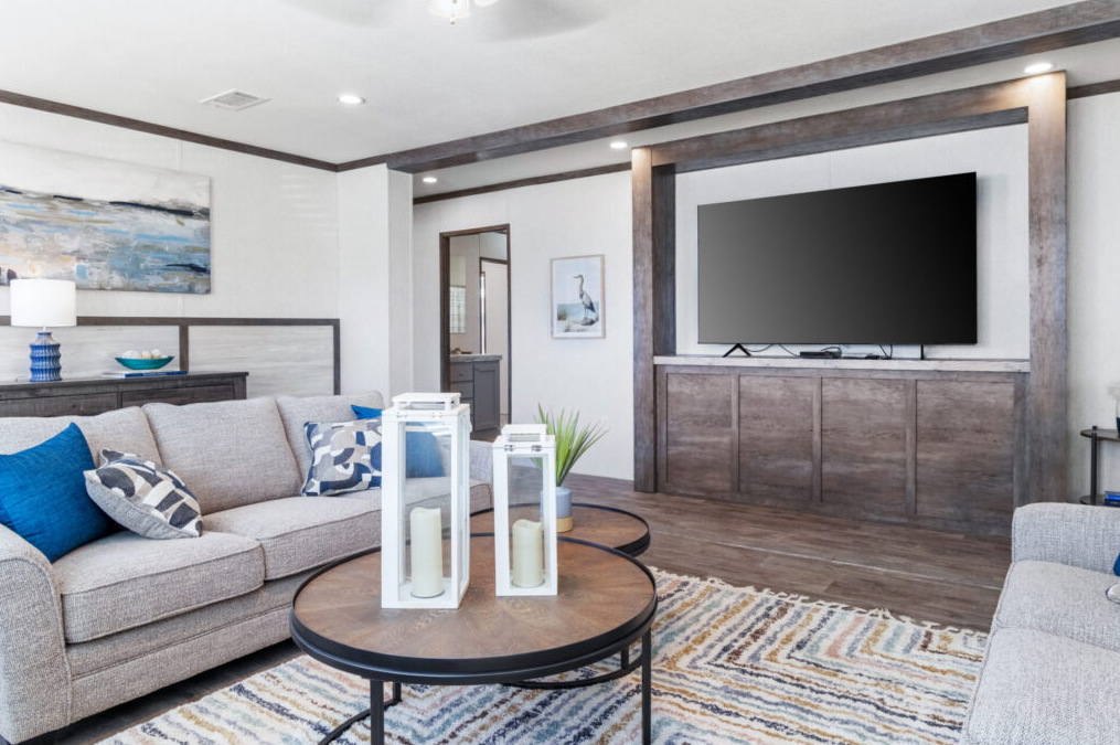 Living room with a beige sofa, blue throw pillows, wooden coffee table with white candles and lanterns, wall-mounted flat-screen TV, artwork, and a patterned area rug.