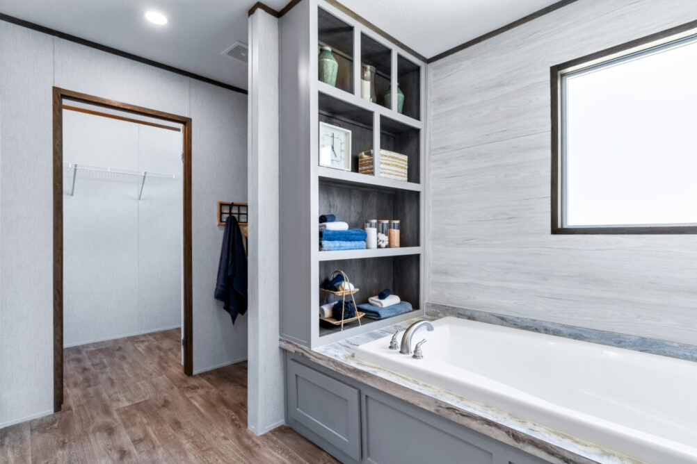 Bathroom with a built-in bathtub, open shelving with towels, decor, and toiletries, window, and a walk-in closet with a hanger and hanging clothes.