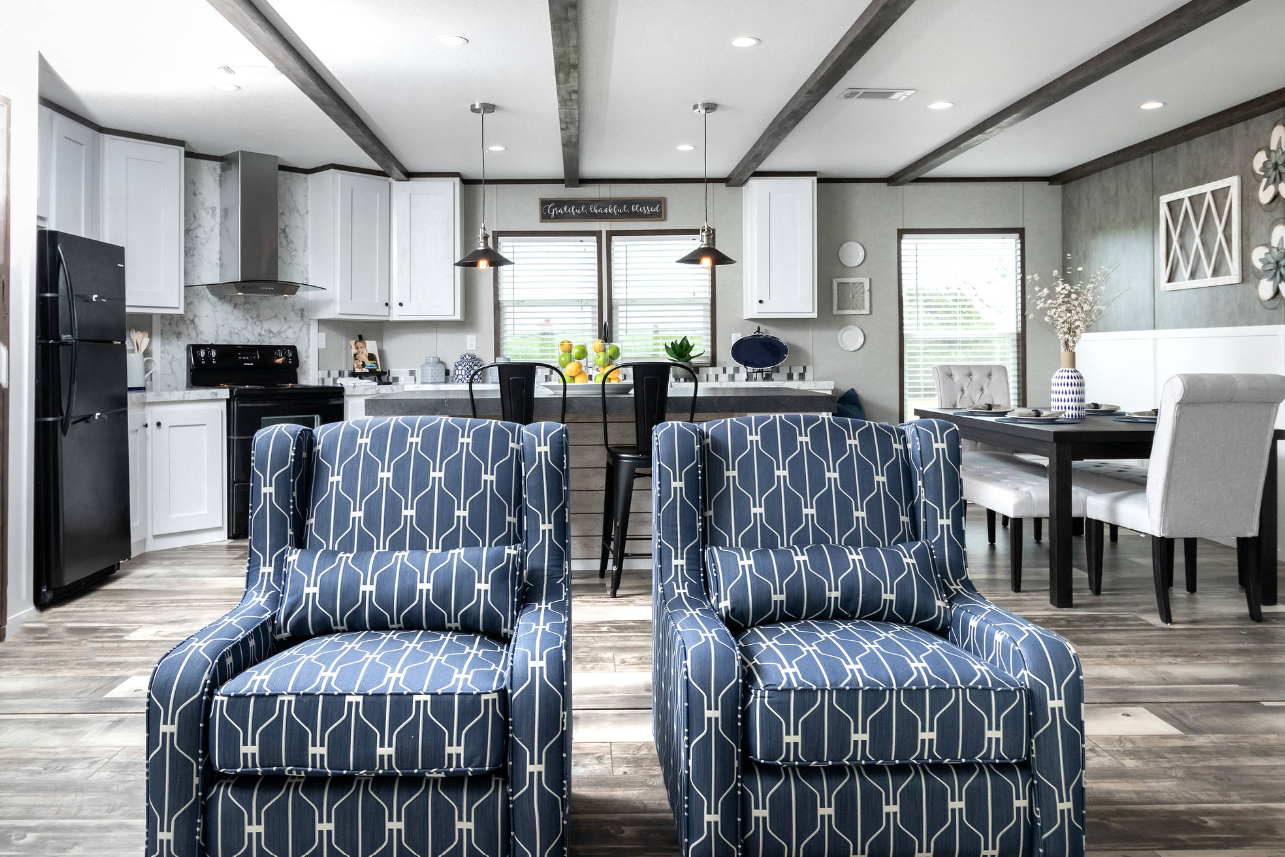 Open-concept living room and kitchen with white cabinets, black appliances, blue patterned armchairs, a dining area with a white table and beige chairs, and decorative accents.
