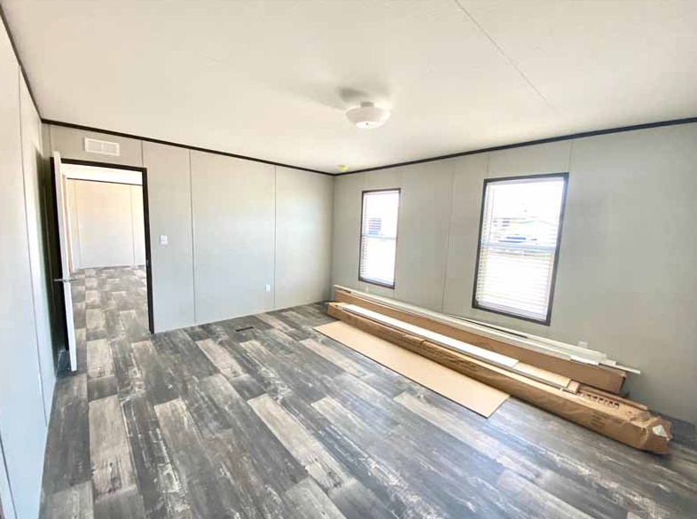 Empty room with gray walls, dark wood flooring, two windows with blinds, and a ceiling light fixture. There are long cardboard boxes and baseboards on the floor near the wall.
