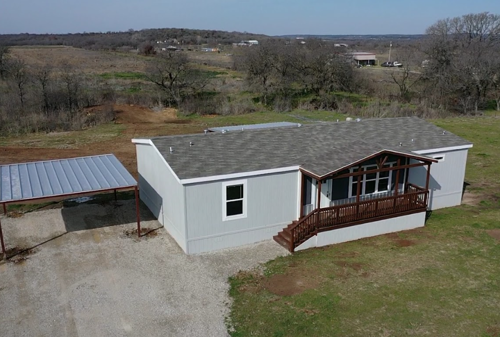 A single-story light gray house with a wooden porch and stairs, situated on a grassy lot with a gravel driveway. There is a small metal-roofed carport on the left side of the house, providing optimal investment opportunity with low barrier to entry