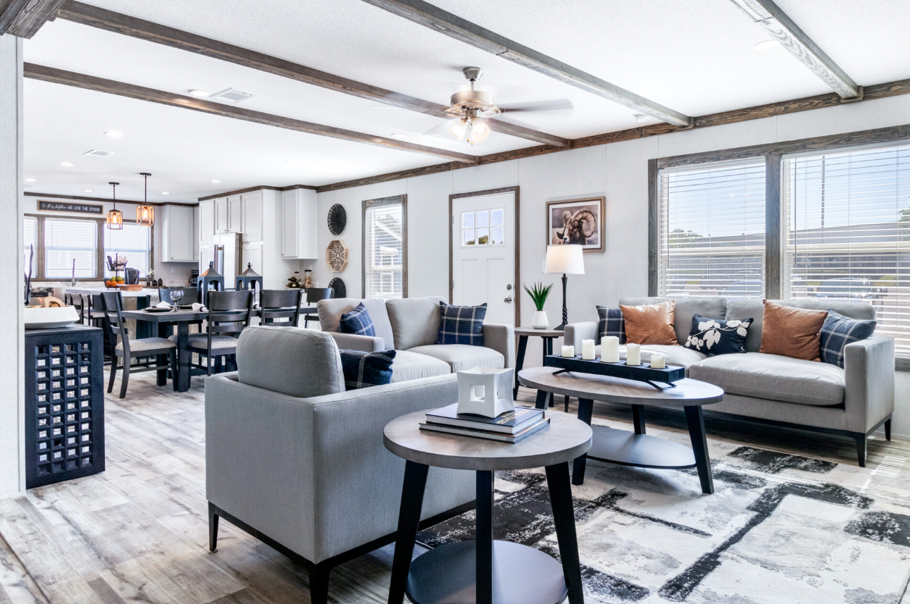 Open-concept living room and dining area with neutral-colored sofas, a wooden coffee table with candles, a side table with a lamp, and a dining table with chairs, featuring large windows with blinds, wood accents on the ceiling, and a kitchen in the 