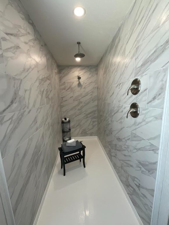 Bright, spacious shower with marble tile walls, two showerheads, a small black bench with folded towels, and a black shelf with shower essentials.