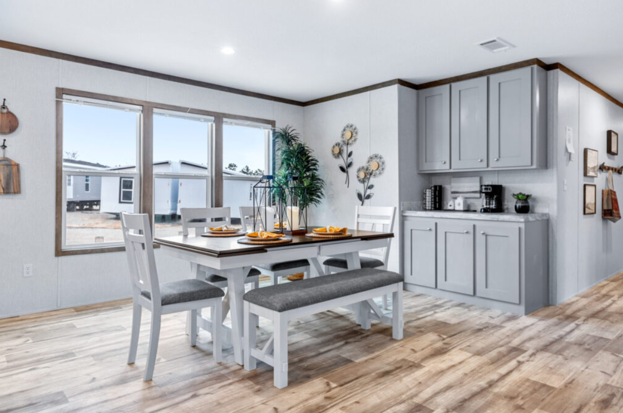 Dining area with a wooden table set for four, gray cushioned chairs, large windows, a gray kitchen cabinet, wall decorations, a potted plant, and a light wooden floor.