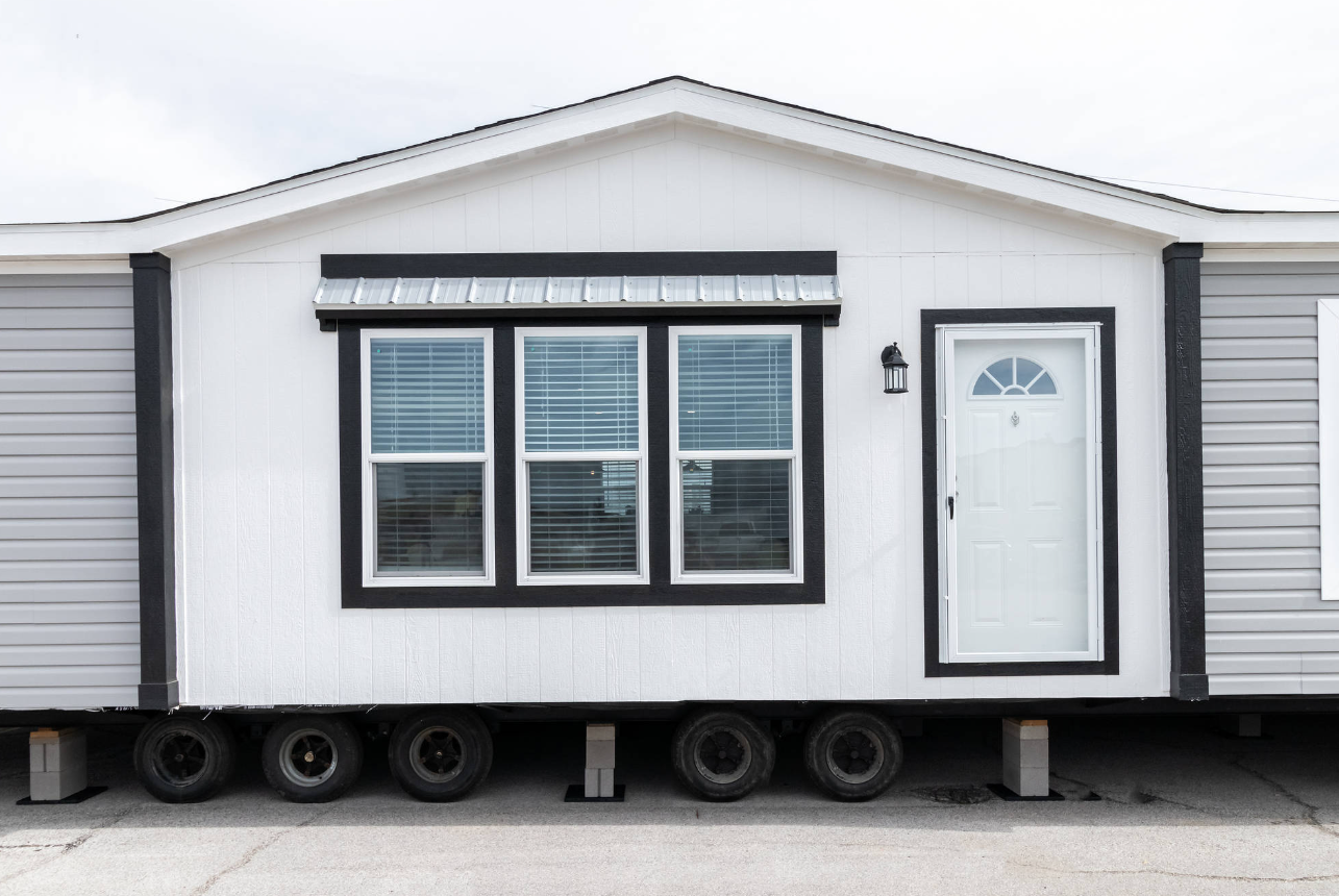 A tiny house on wheels with white siding, black trim, three large windows with blinds, a white front door with a semi-circular window, a black wall-mounted lantern, and a metal awning above the windows. The house is elevated on blocks and tires.