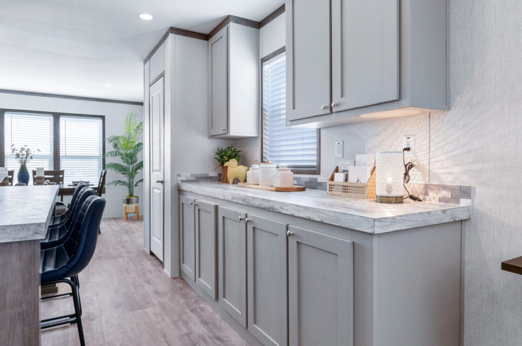 Kitchen with light gray cabinets, a marble countertop, and a window with blinds. Dining area with chairs and large windows in the background.