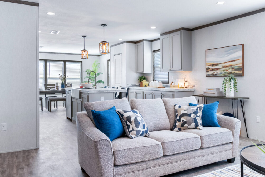 Living room with a gray sofa, blue and patterned throw pillows, open kitchen and dining area, large windows with blinds, wall art, and minimal decor.