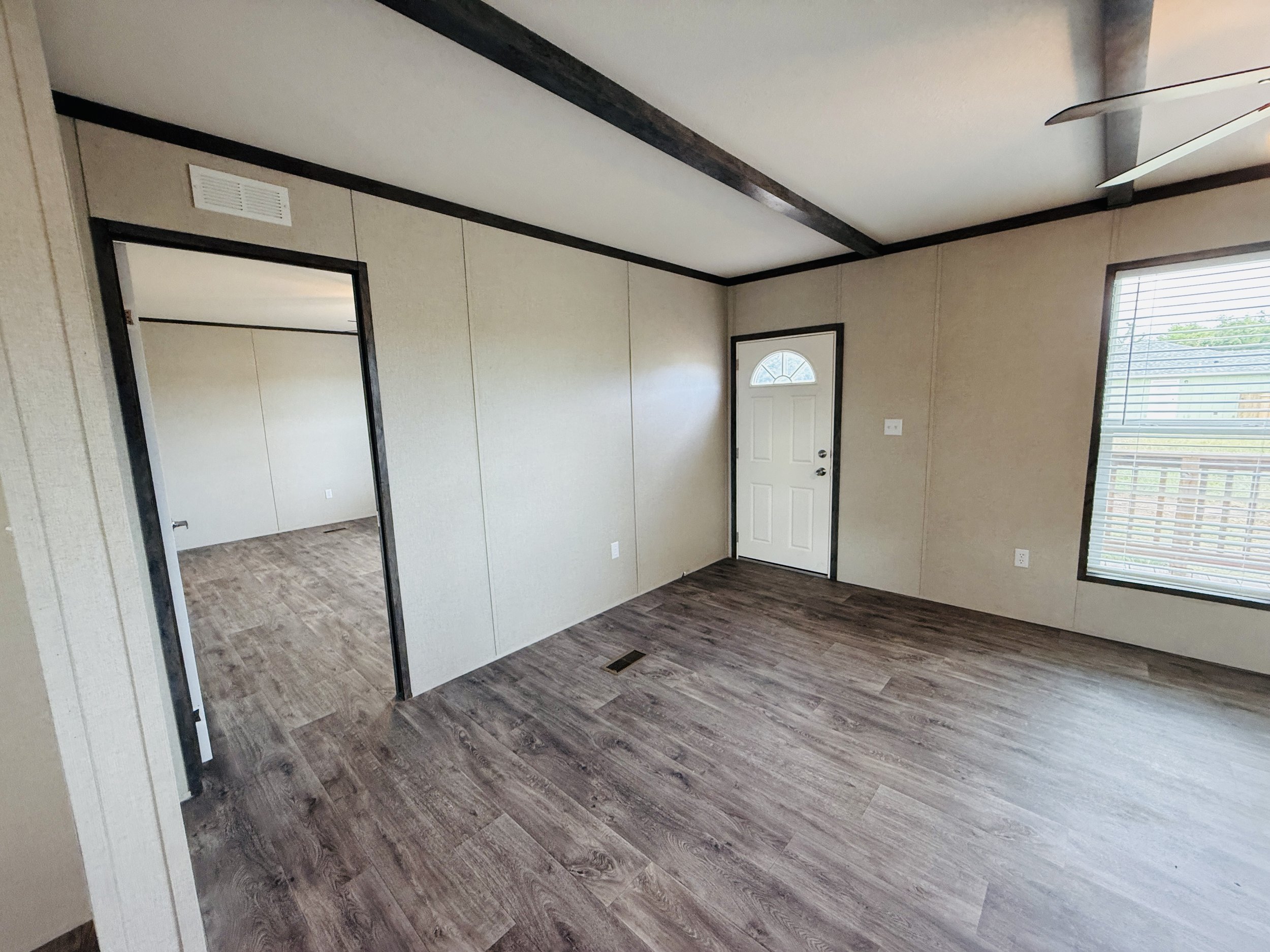 Empty living room with beige walls, dark wood trim, a white door with an arched window, a large window with blinds, wood laminate flooring, and a ceiling fan.