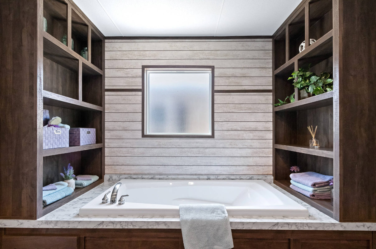 A bathroom with a built-in bathtub surrounded by dark wooden shelves on both sides, a marble countertop, a window above the tub, and decorative items including towels, plants, and candles.