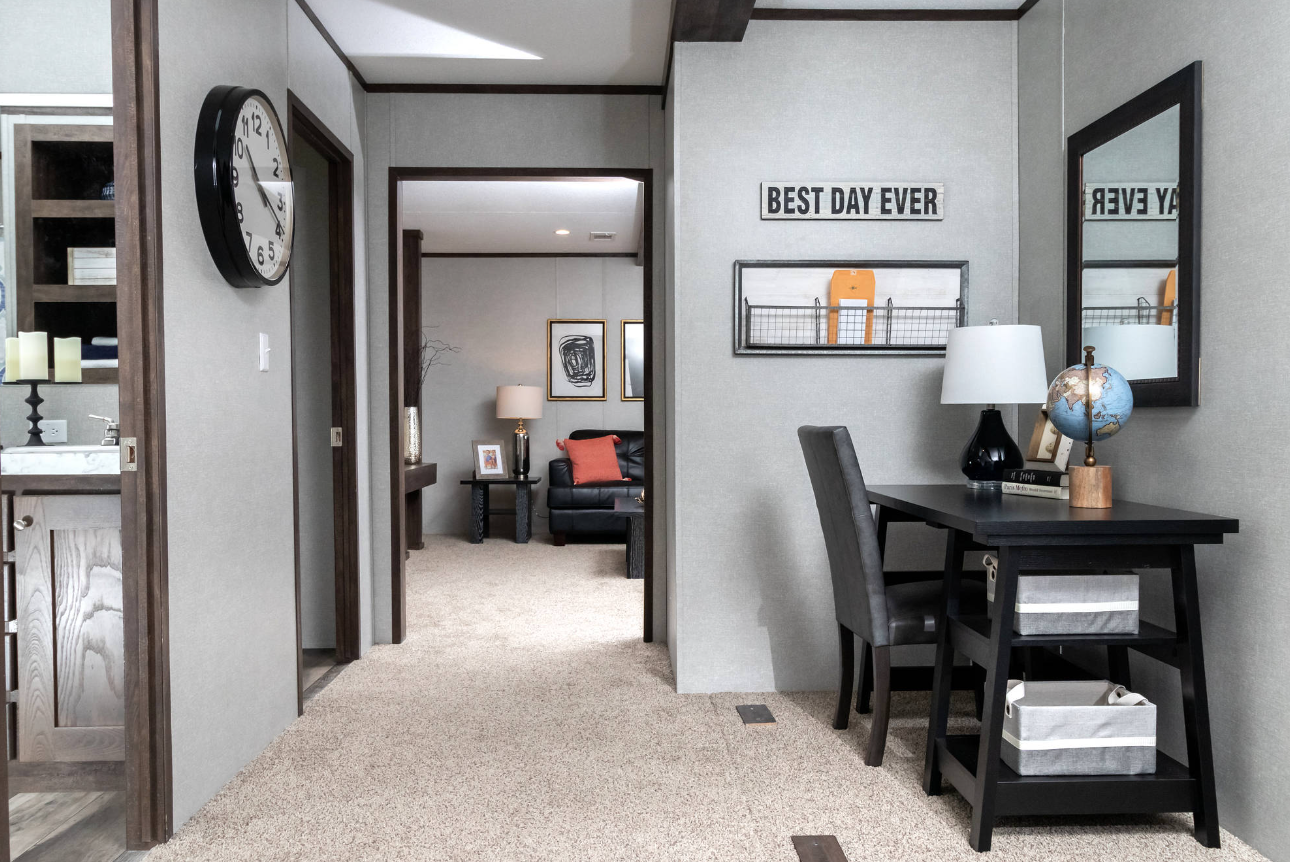 A hallway in a house with a black desk, a chair, a lamp, a globe, and a mirror on the wall. A sign reads "Best Day Ever," and a basket is on the bottom of the desk. There's an open doorway leading to a living room with a sofa and a side table with a 