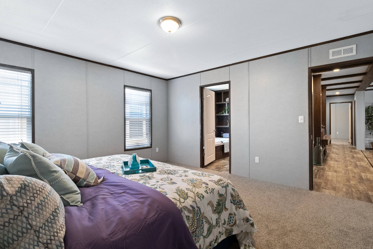 Bedroom with four pillows, floral bedspread, two windows with blinds, door leading to a bathroom, hallway with wooden flooring, ceiling light, and a small tray with teal bottles on the bed.