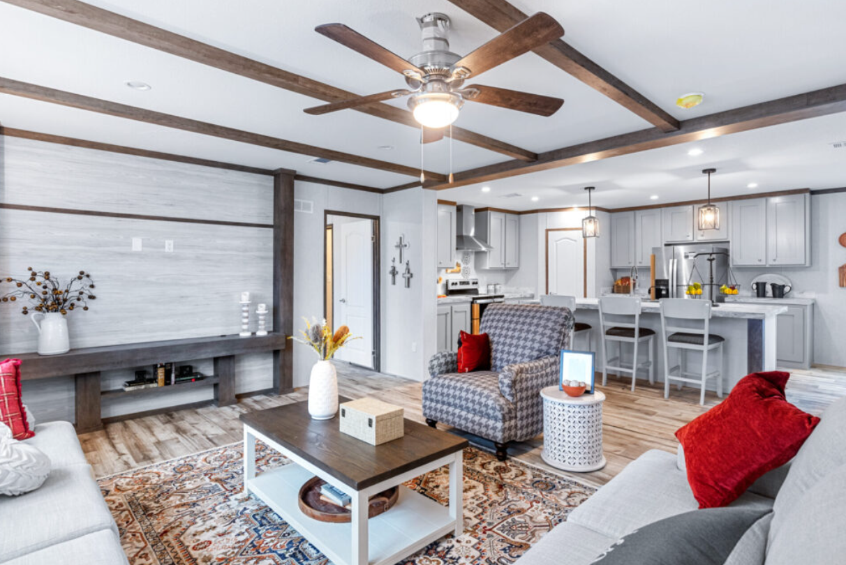 Open-concept living room and kitchen with white and gray decor, wooden accents, ceiling fan, and upholstered furniture with red pillows.