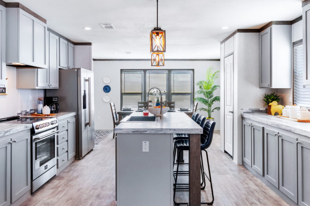 Modern kitchen with light gray cabinets, a center island with bar stools, stainless steel appliances, and large windows with blinds, decorated with potted plants and wall art.