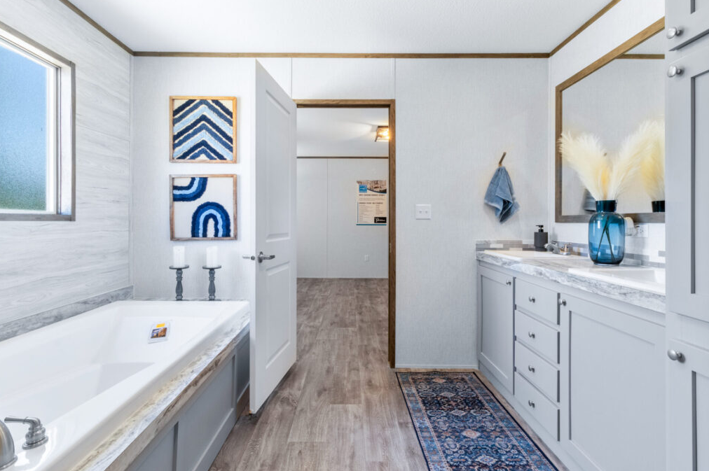 A modern bathroom with a large bathtub, a window, and a vanity with a mirror. The decor features blue and white accents, a framed art three pieces on the wall, a blue vase with pampas grass, and a blue towel hanging on the wall.