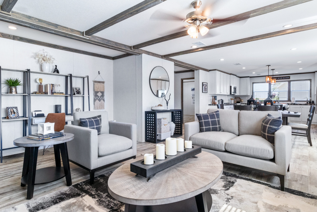 Living room with light-colored couches, a black coffee table with candles, a bookshelf, and a view of the open kitchen in the background, decorated in modern style with neutral tones and wood accents.