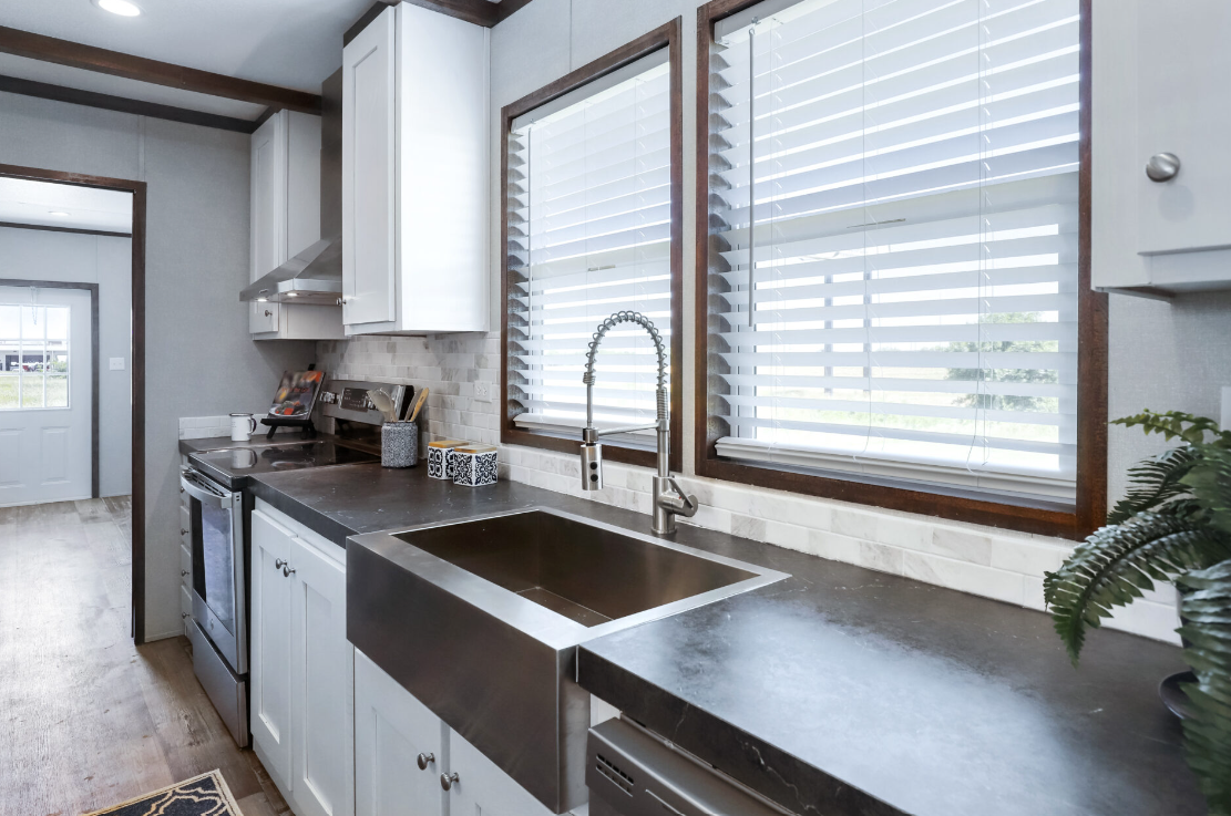 Kitchen with white cabinets, a large stainless steel farm sink, a window with blinds above the sink, and a black countertop. A stove, some small kitchen items, and a potted plant are also visible.
