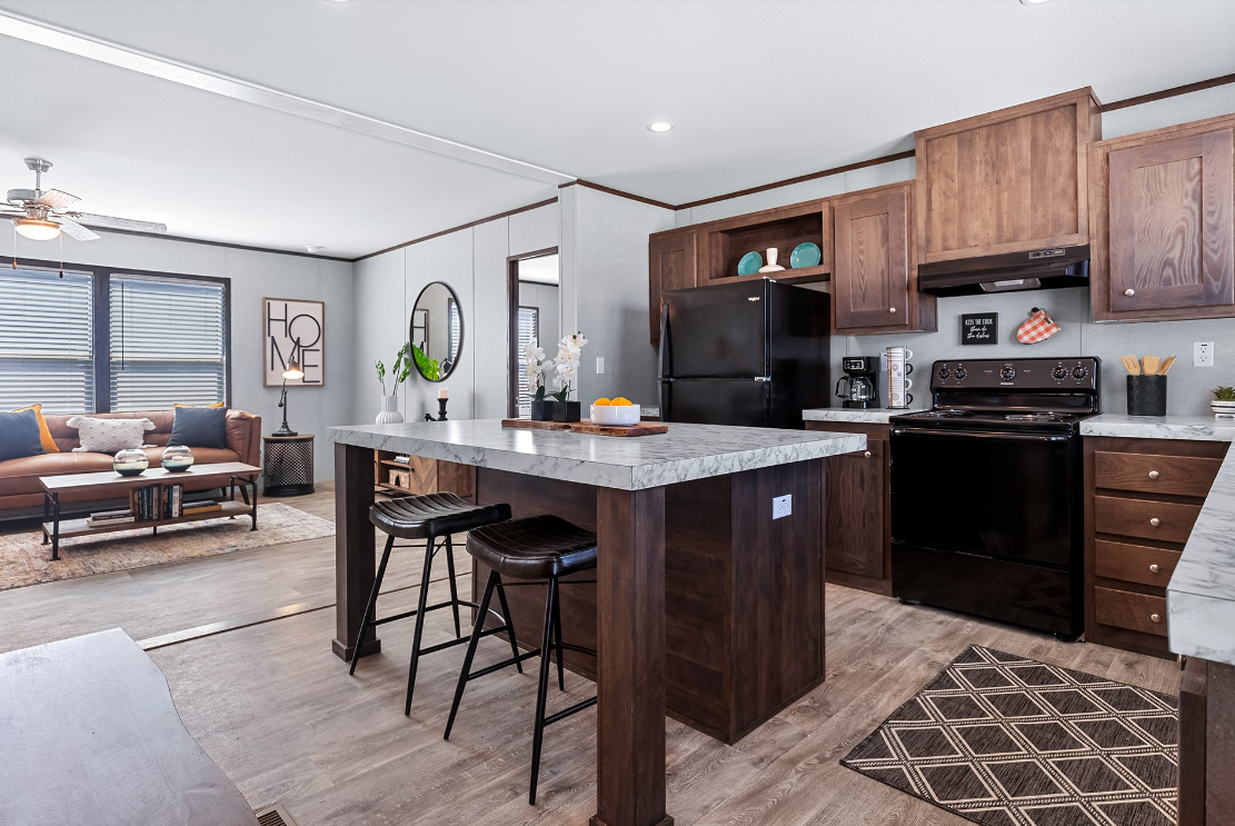 Open-concept living room and kitchen with wooden cabinets, a black refrigerator, and a marble kitchen island with two black barstools. The living room has a brown sofa with navy and beige pillows, a wooden coffee table, and large windows with blinds.