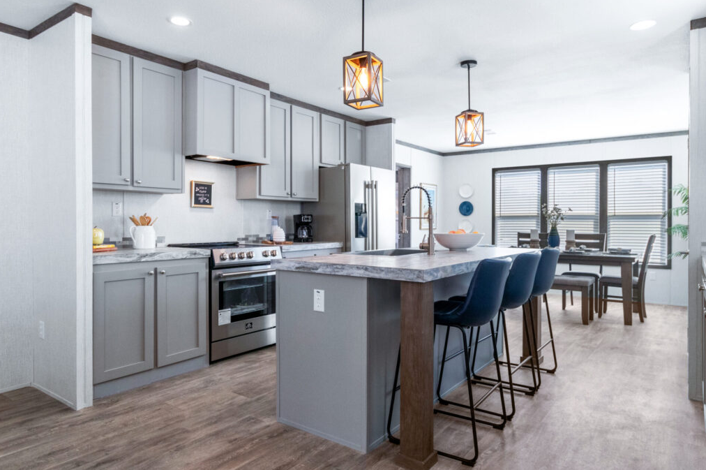 Modern kitchen with gray cabinets, stainless steel appliances, a marble island with blue bar stools, and a dining area with a table and chairs near large windows.