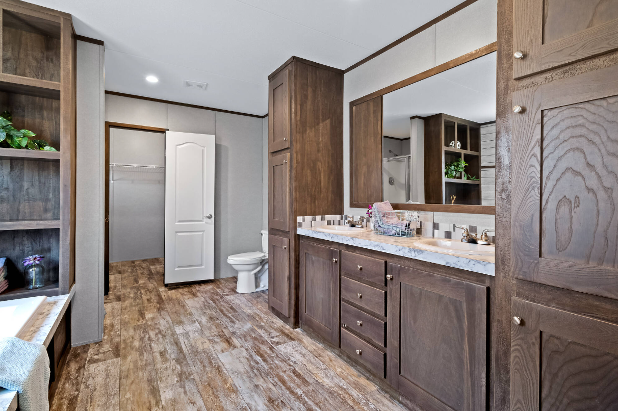 Bathroom with wooden cabinetry, a large mirror, dual sinks, and a marble countertop. There is a laundry closet, a toilet, shelves with plants and decor, and wood-look flooring.