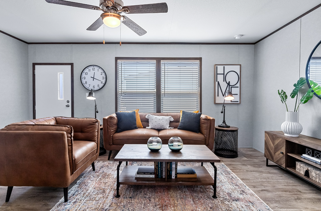 Living room with gray walls, a ceiling fan, large window with blinds, brown leather sofas, a wooden coffee table with decorative glass bowls, a patterned rug, wall clock, and framed artwork that says 'HOME'.
