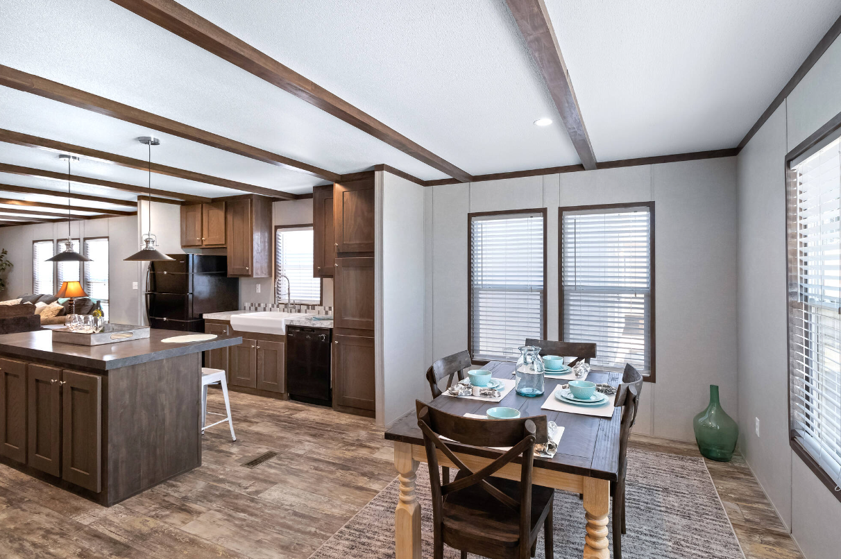 Interior view of a kitchen and dining area in a home with wood floors, white walls, and wooden beams on the ceiling. The kitchen has dark wood cabinets, a white sink, black refrigerator, and black dishwasher. The dining area has a wooden table set wi