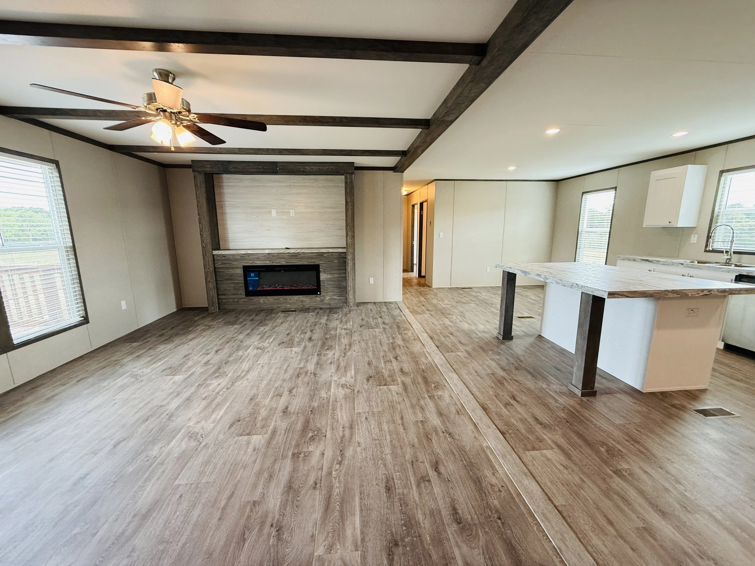 Open living room and kitchen with wood floors, a ceiling fan, a fireplace, large windows, and a kitchen island with a marble countertop.