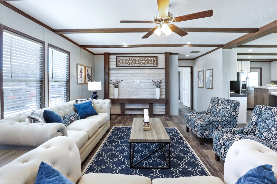 Living room with white and patterned sofas, blue pillows, wooden coffee table, navy rug, large windows with blinds, ceiling fan, and wall art.