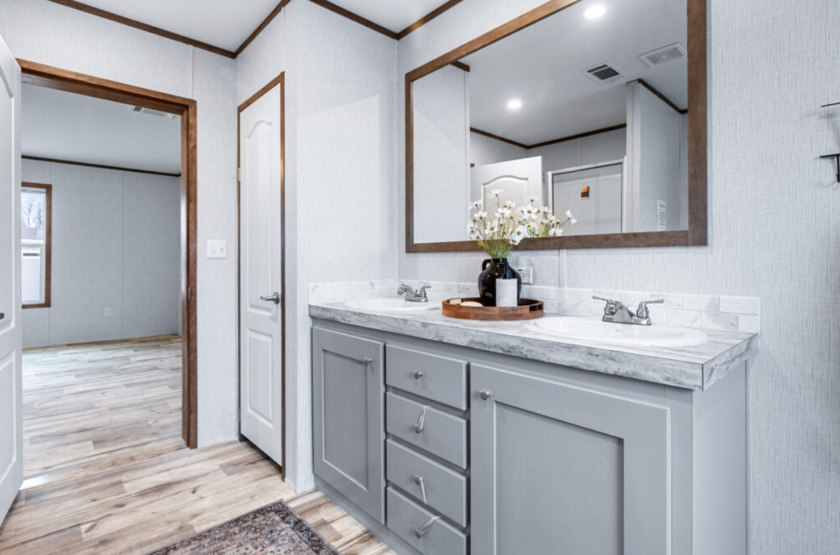 Bathroom vanity with two sinks, a large mirror, and glossy gray cabinets, with a tray holding a black vase and flowers on the countertop.
