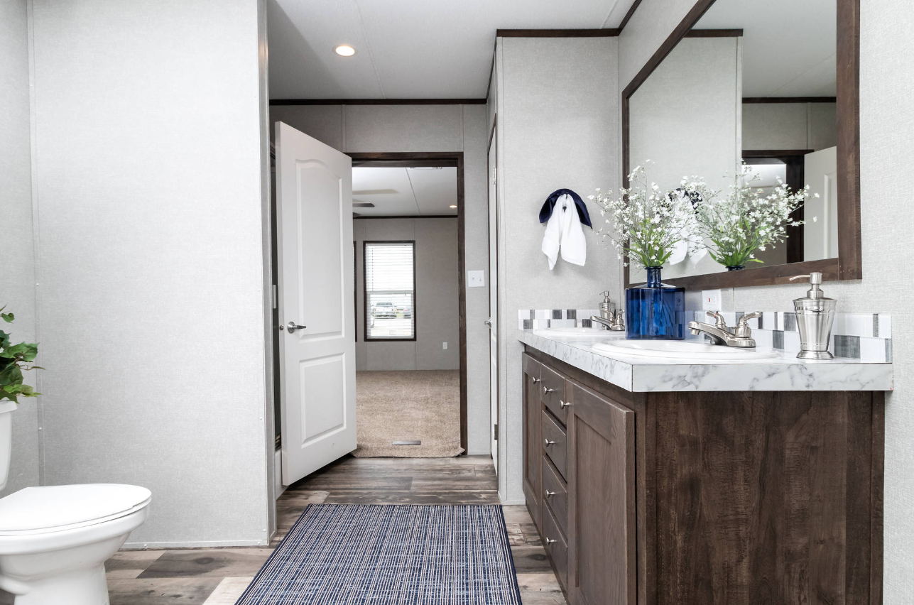 Bathroom with dual sinks, large mirror, flower vase, towel, and view into a carpeted room with a window.