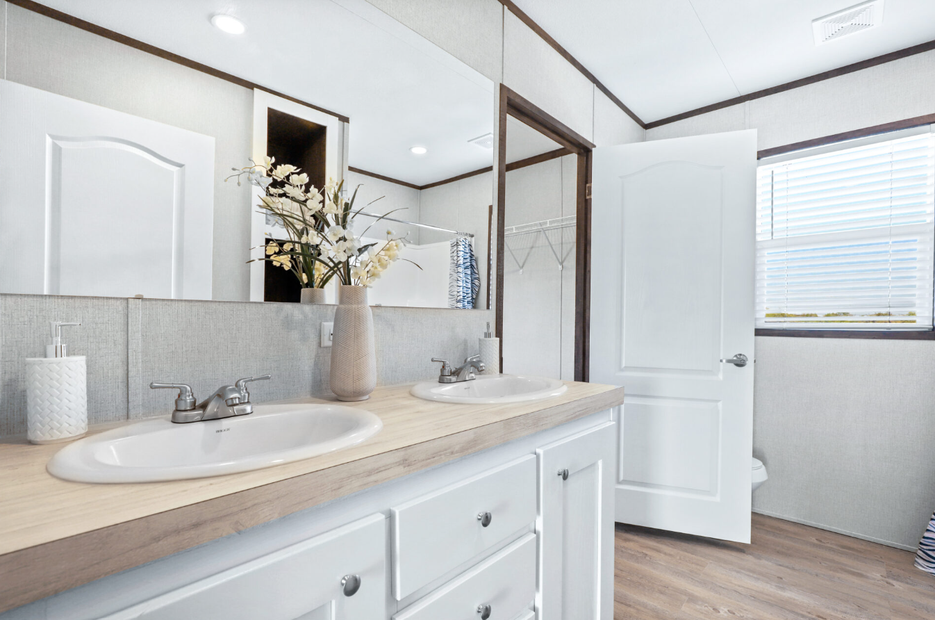 A bathroom with a double sink vanity, large mirror, white cabinets, a vase with flowers, soap dispenser, and a window with blinds.