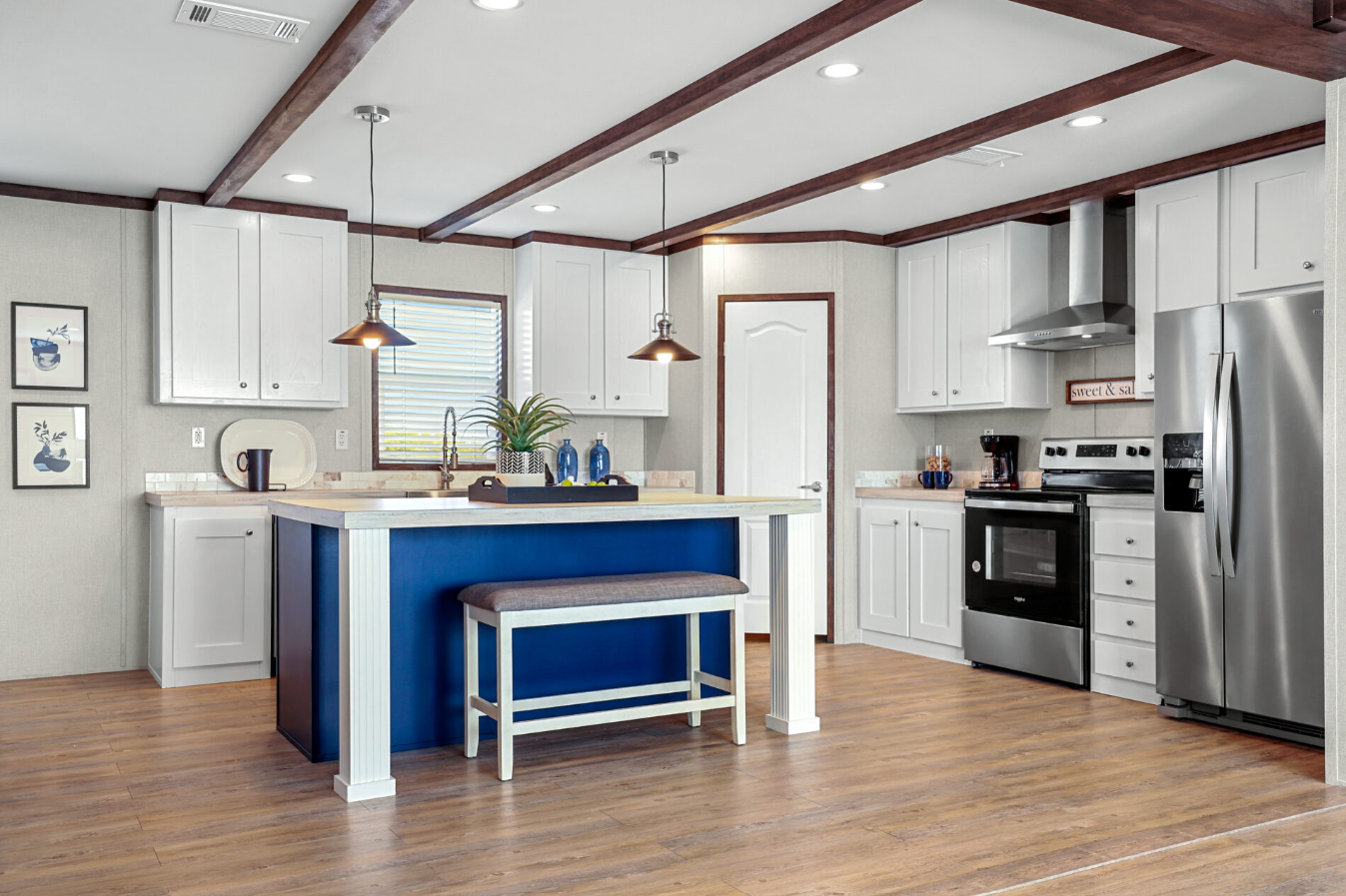 Modern kitchen with white cabinets, stainless steel appliances, a blue island with a bench, hardwood flooring, pendant lights, window with blinds, and wall art.
