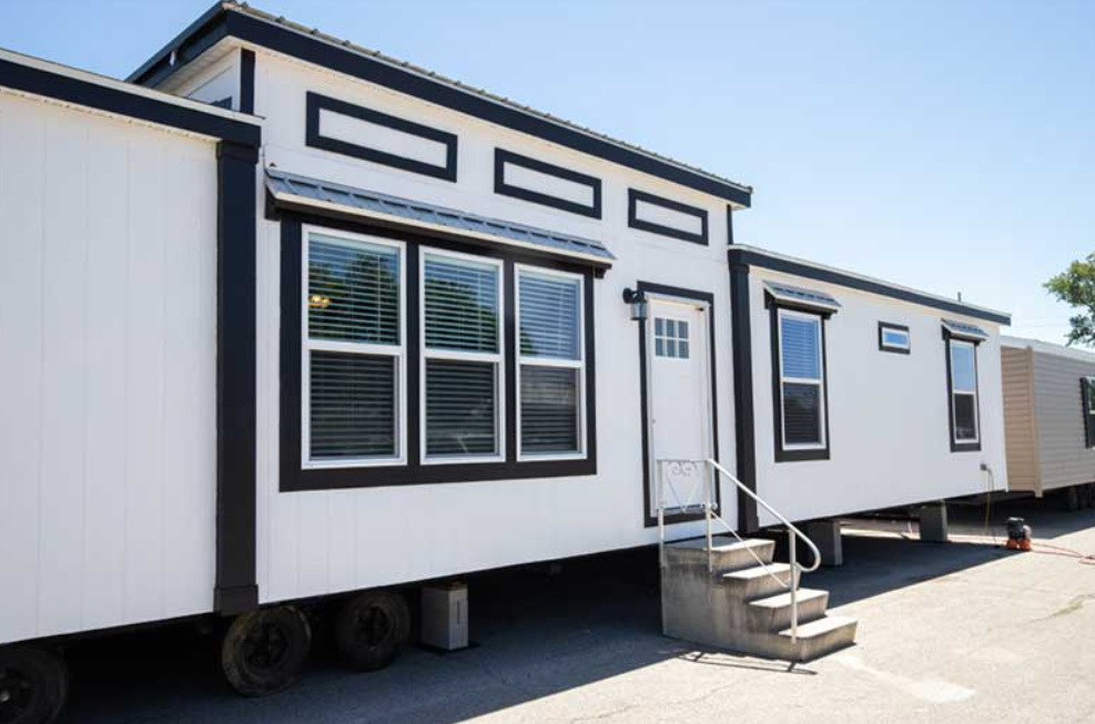 White manufactured home with black trim and multiple windows, including a small door with steps, standing on wheels in an outdoor lot.