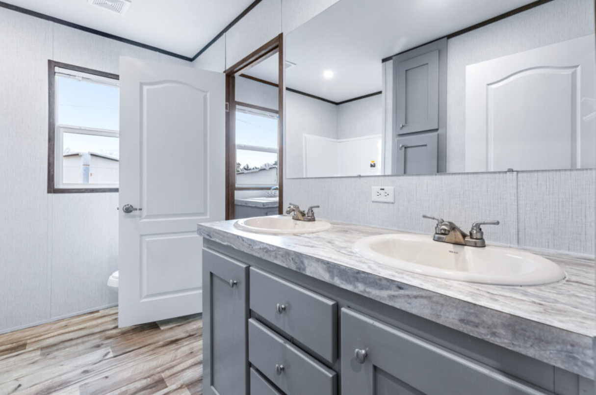 Bathroom with a double sink vanity, gray cabinets, marble countertop, large mirror, white door, and wood-style flooring.