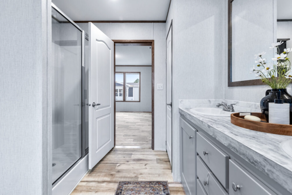 Bathroom with a vanity, marble countertop, sink, mirror, flowers in a black vase, soap on a wooden tray, as seen from the hallway into the adjacent room with windows.