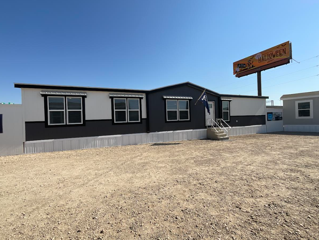 A manufactured home with black and white exterior paint, four windows, a small set of stairs with a flag. Providing optimal investment opportunity with low barrier to entry and high-yield returns.