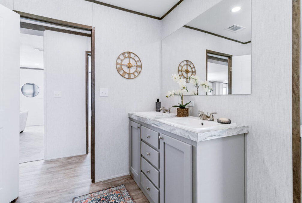 Bathroom vanity with a white marble countertop, a large mirror, a vase with white orchids, and a wall clock, with a doorway leading to a living room in the background.
