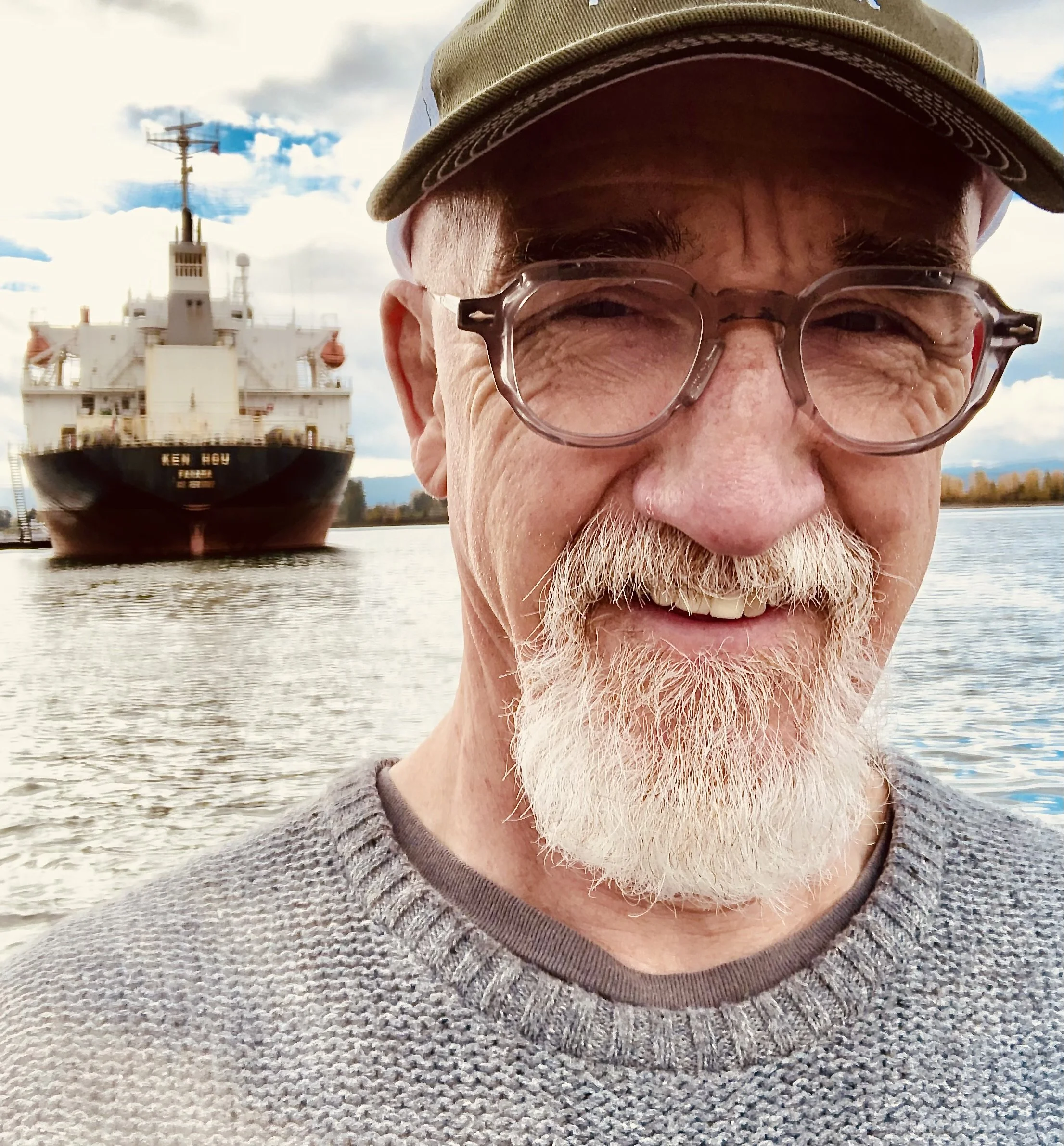 A close-up of a man smiling, wearing glasses, a tan cap, and a gray sweater. Behind him, a large ship is visible on the water with a partly cloudy sky above.