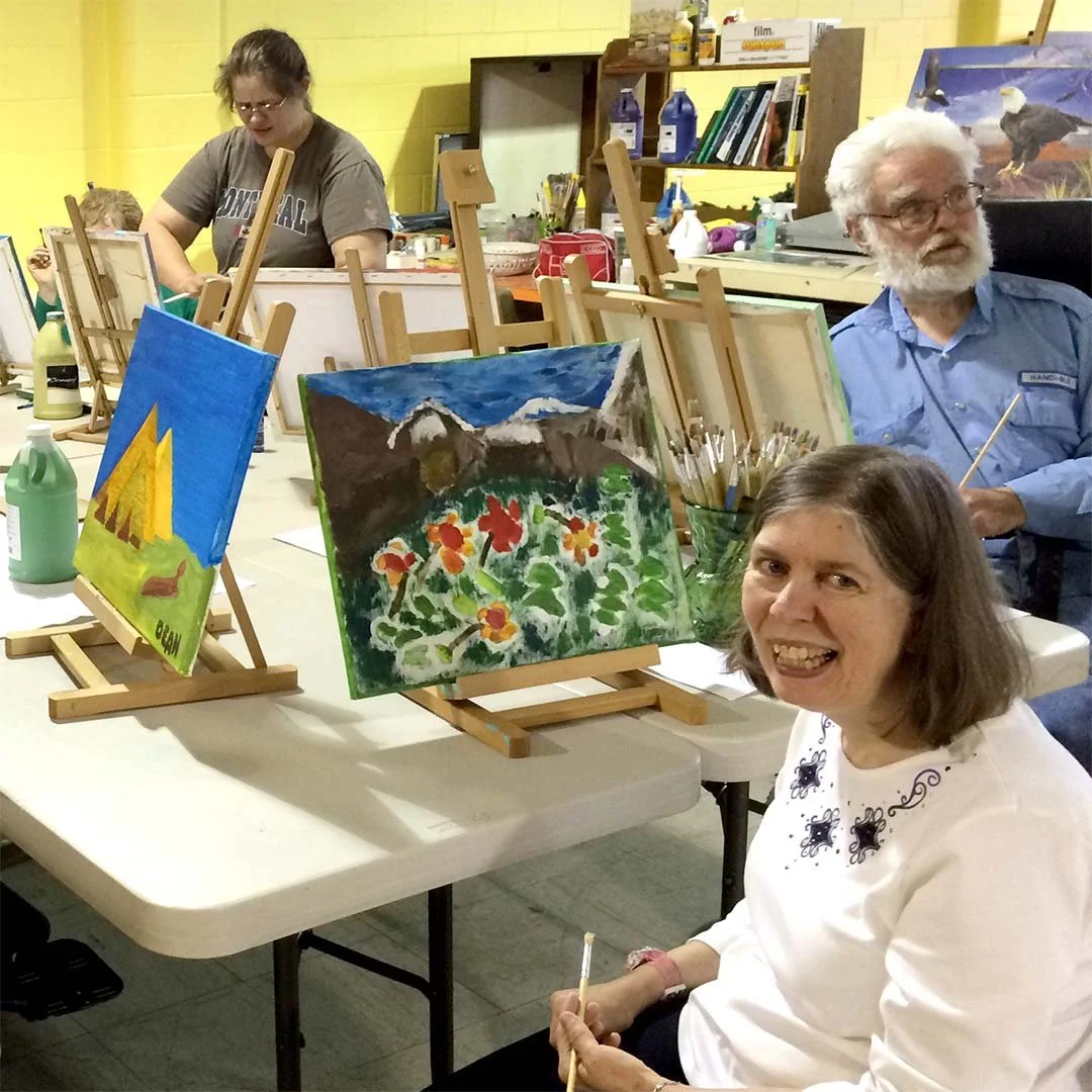 A community member takes a break from painting to smile at the camera with other members painting in the background during an art project