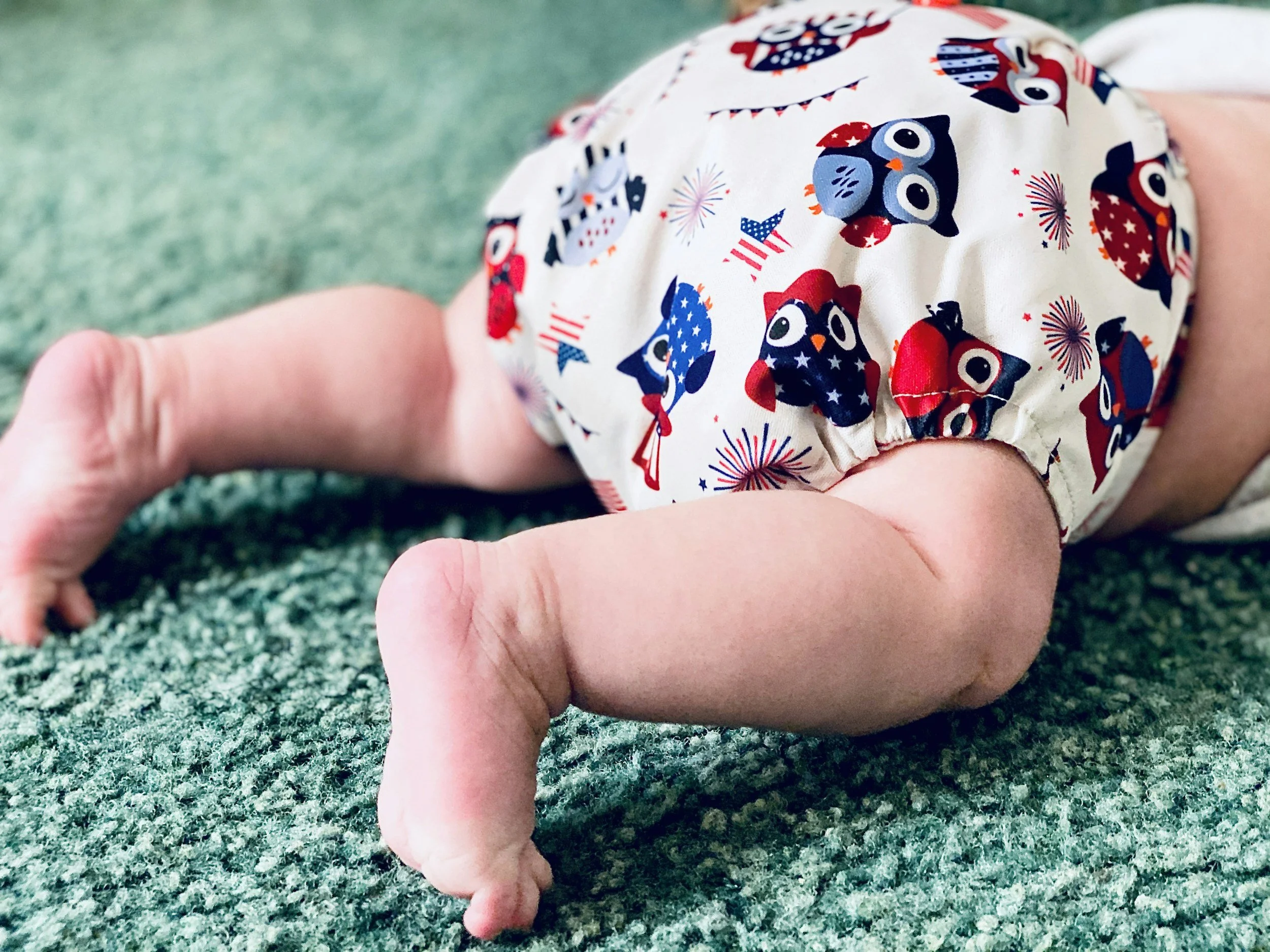 A baby crawling on a green textured carpet, wearing a diaper with patriotic owl and fireworks pattern in red, white, and blue.